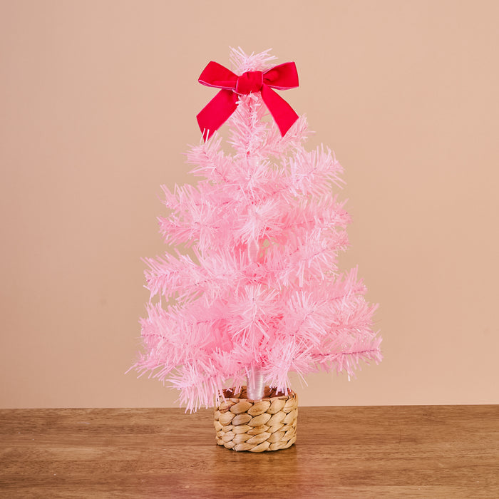 A small, bright pink Pink Mini Artificial Christmas Tree from The Darling Effect, featuring a red bow at the top and a woven basket base for holiday décor.