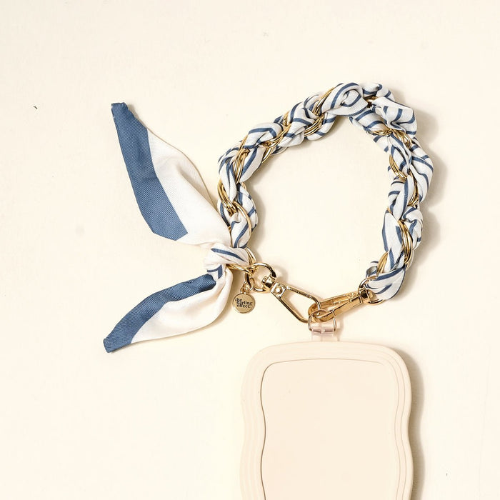 The Darling Effect’s Bluebell Stripe Call Me Later Phone Scarf features a braided blue, white, and gold ribbon with gold-tone chain hardware, a heart charm, and a cream wavy-edged tag, displayed on a light background.