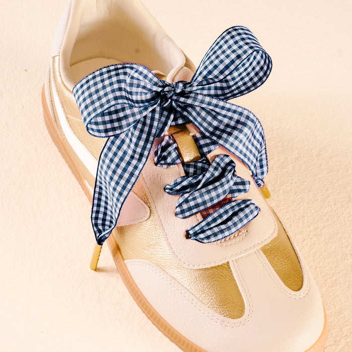 The Darling Effect's Gingham Navy Step It Up Shoelaces, featuring a large blue and white gingham bow, are shown laced on a white and gold sneaker against a light beige background.