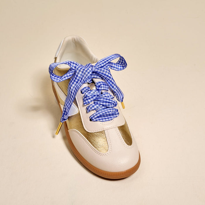 The Darling Effect’s Gingham Royal Blue Step It Up Shoelaces—blue and white checkered, durable, and tied in a bow—adorn a white and gold sneaker with tan soles, shown on a light beige background.