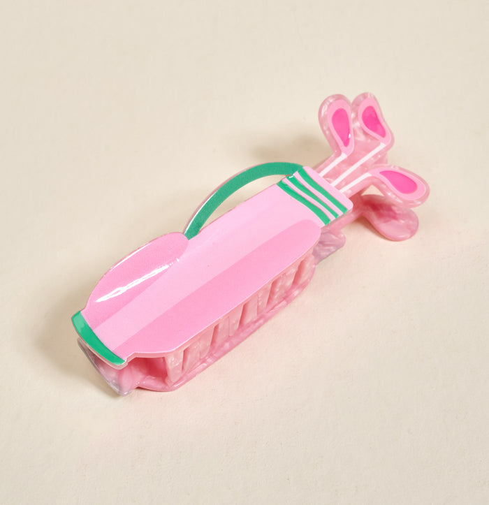 The Darling Effect’s Golf Clubs Shape Up Claw Clip is a playful pink plastic hair accessory shaped like a golf bag with green, white, and pink club details—perfect for cute hairstyles. Shown on a light beige surface.