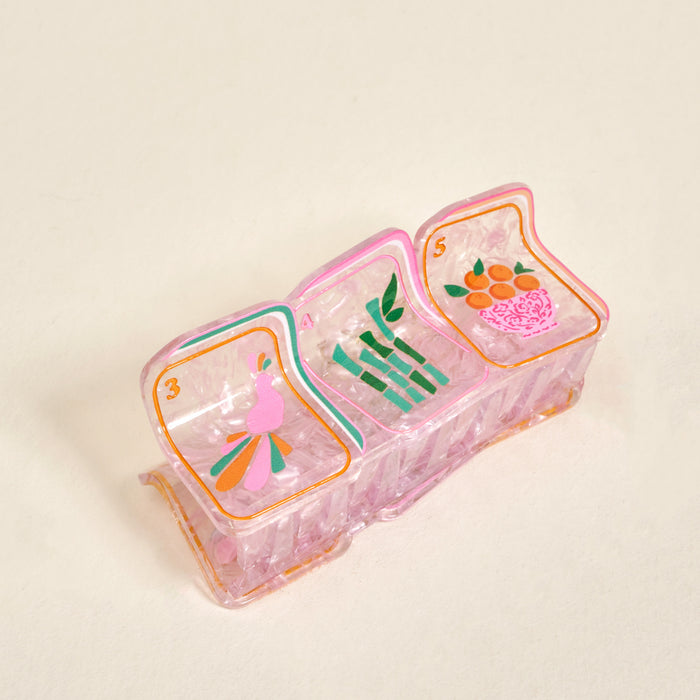 A vintage pink plastic divided tray featuring whimsical cartoon art of The Darling Effect’s Mahjong Tiles Shape Up Claw Clip, a pink bird, bamboo stalks, and oranges in a basket.