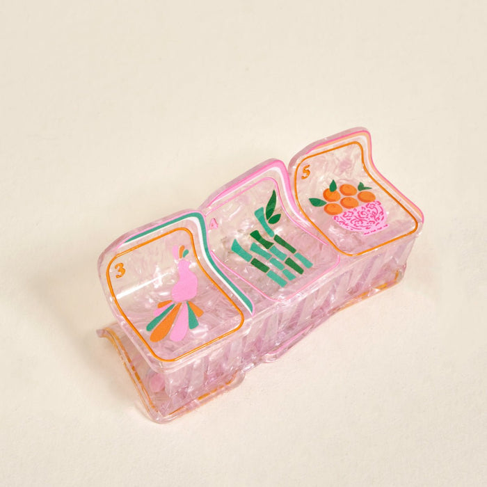 A vintage pink plastic divided tray featuring whimsical cartoon art of The Darling Effect’s Mahjong Tiles Shape Up Claw Clip, a pink bird, bamboo stalks, and oranges in a basket.