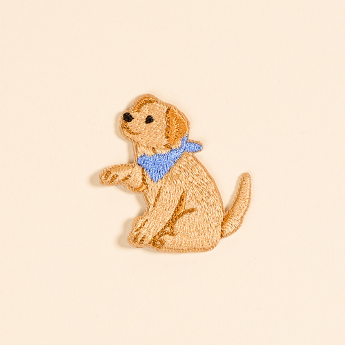 An embroidered patch of a light brown dog sitting and wearing a blue bandana, shown on a plain beige background.