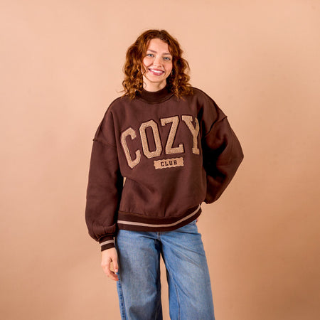 A smiling person with curly hair wears The Darling Effect’s Cozy Club Mockneck Sweatshirt in brown paired with blue jeans, standing against a plain tan background and showcasing the relaxed, cozy comfort of its silhouette.
