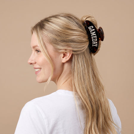 A woman with long blonde hair in a white shirt smiles, her hair clipped up with a large brown hair clip that says GAMEDAY in white letters. The background is plain beige.