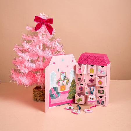A pink, house-shaped Adoring Ornaments Ornament Advent Calendar with twelve numbered drawers stands next to a small pink artificial Christmas tree, with miniature acrylic ornaments spread on the surface in front.