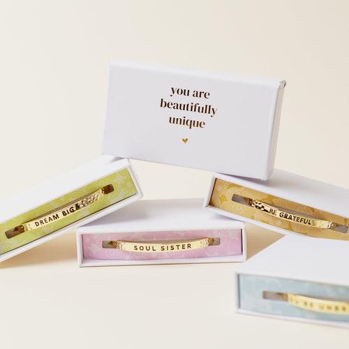Four gold bracelets in colorful boxes, each engraved with positive messages: DREAM BIG, SOUL SISTER, BE GRATEFUL. A white box above reads, you are beautifully unique.