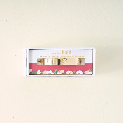 A gold bracelet in a white box with a card that says You are bold. Love who you are and forget what others think. The box and card have floral designs on the bottom.