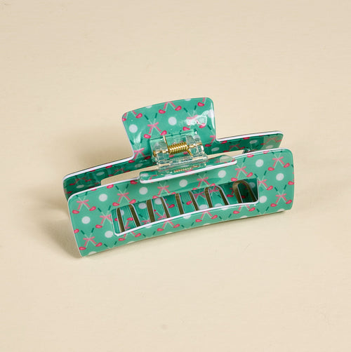 A large teal hair claw clip with white polka dots and pink X patterns is shown open on a plain beige surface.