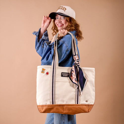 A smiling person and The Darling Effect’s Gameday Classic Canvas Hat holds a large beige tote bag with brown trim, striped handles, and a scarf tied on. The bag is decorated with patches and pins. The background is a solid light brown color.