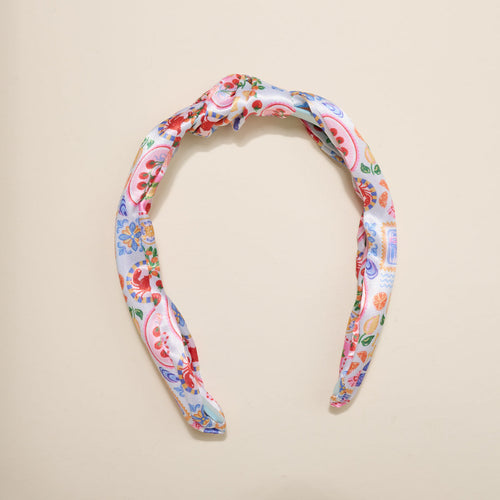 A fabric headband with a knot at the top, featuring a colorful floral and paisley pattern on a white background, displayed on a neutral surface.