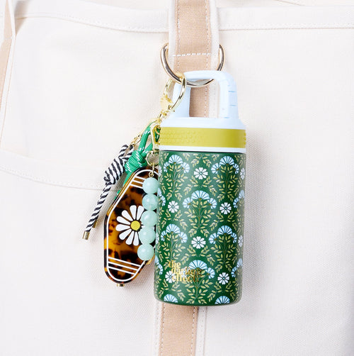 The Darling Effect’s Everbloom Forest Green 6 oz Lil' Bitty Bottle, with a yellow cap and green floral pattern, hangs from a beige bag by carabiner next to a beaded keychain—a stylish hand sanitizer ideal for travel.