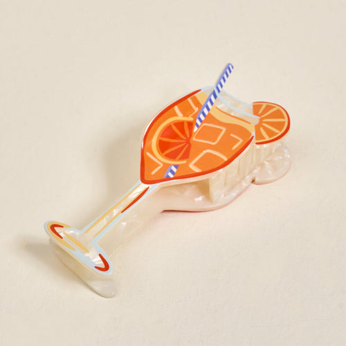 A colorful hair clip shaped like an orange cocktail glass with ice cubes, a straw, and an orange slice, resting on a light beige surface.