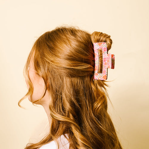 A woman with wavy, light brown hair is shown from the side, wearing a large pink floral hair clip against a light beige background.