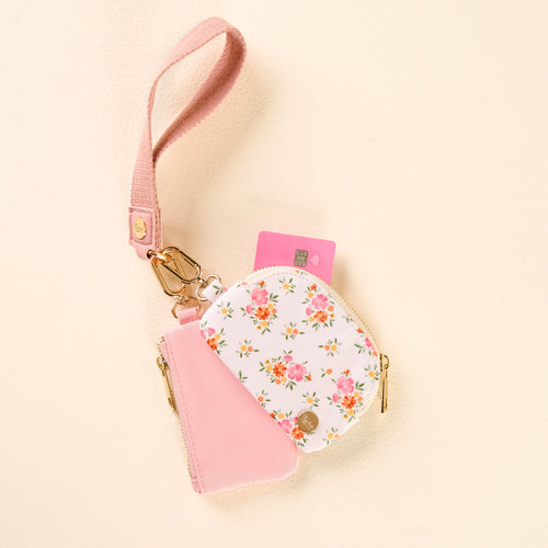 A small floral and pink coin purse with a wrist strap, holding a pink card and a zipper pocket, displayed on a light cream background.