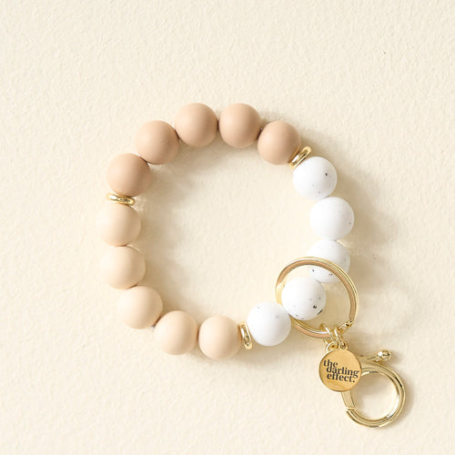 A beaded keychain bracelet with tan and white round beads, gold rings, and a gold charm engraved with The Giving Effect, displayed on a cream-colored surface.