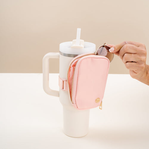 A hand places round sunglasses into The Darling Effect's Dusty Blush Tumbler Fanny Pack, shown attached to a stylish white tumbler with straw and handle, all set against a light beige background.