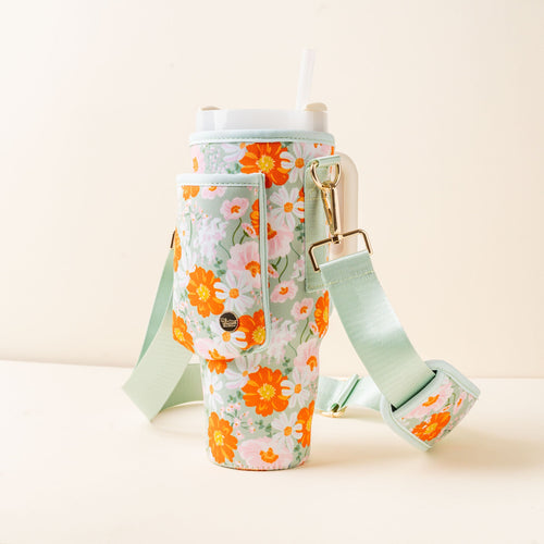 The-darling-effect's Bouquet Beauty Sage Tumbler Sling includes a tall tumbler with lid, straw, and a pastel floral sleeve with orange, yellow, and white flowers. The sling has a pocket, snap button, and adjustable strap for hands-free carrying.