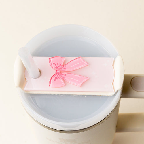 A top-down view of a cup with a lid, featuring a pink bow decoration and a straw inserted through the lid. The cup is filled with a light-colored liquid.