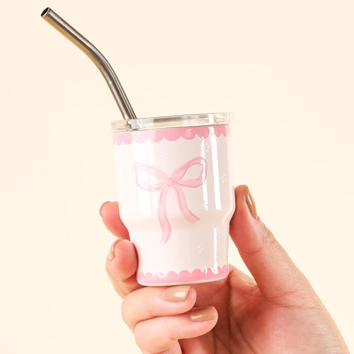 A hand holds a white cup with pink scalloped edges and a pink bow design. The cup has a clear lid and a bent metal straw. The nails are painted beige. The background is light cream.