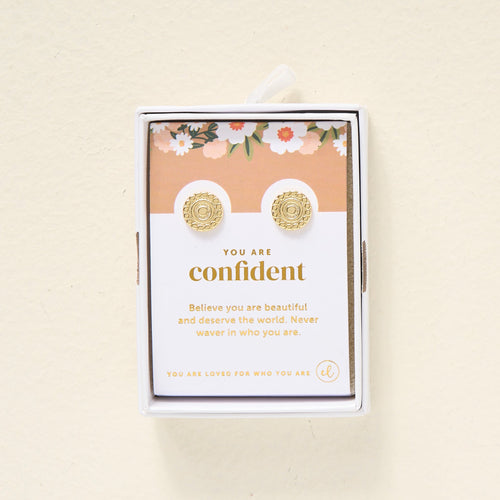 A pair of gold stud earrings displayed on a card that reads, You are confident. Believe you are beautiful and deserve the world. Never waver in who you are.