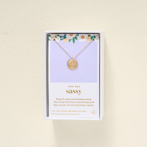 A gold pendant necklace in a white gift box with a floral design and a card that reads You are sassy along with an uplifting message about being quick-witted and dramatic.