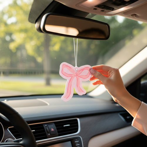 A person hangs a pink bow-shaped air freshener on the rearview mirror inside a car, with sunlight and green trees visible through the window.
