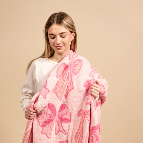 A young woman with straight blonde hair and a white top gently smiles down while holding the-darling-effect’s Blushing Bows Pink Cozy Dreams Luxe Blanket—an ultra-soft throw featuring large pink bows—against a plain beige background.