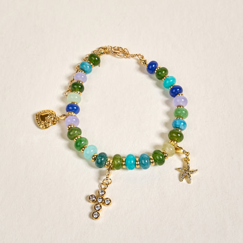 The Darling Effect's True Blues Stone Candy Bracelet showcases blue, green, and turquoise beads with gold accents and three gold charms—a heart, rhinestone cross, and starfish—beautifully arranged on a light, textured surface.