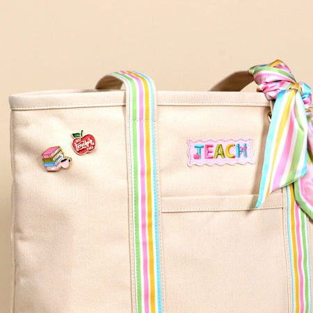 A beige tote bag with pastel rainbow straps, a matching scarf tied to the handle, a “TEACH” patch, a books pin, and a red apple pin that says “teach” on the front pocket.