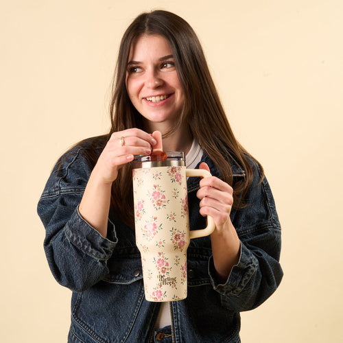 A smiling woman in a denim jacket holds the-darling-effect Endless Daydream Cream 40 oz Lifestyle Flip Straw Tumbler, adjusting its lid with one hand while standing before a plain beige background.
