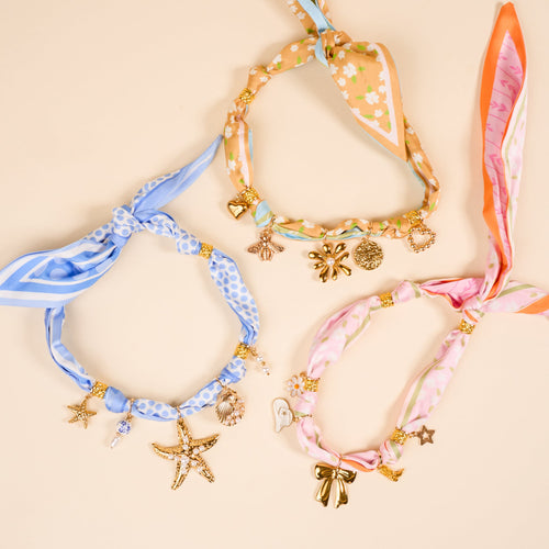 The Darling Effect's Blushing Hour Make a Statement Scarf Necklace features bold blue, peach, and pink patterns—polka dots, florals, stripes—with gold charms, artfully arranged on beige.