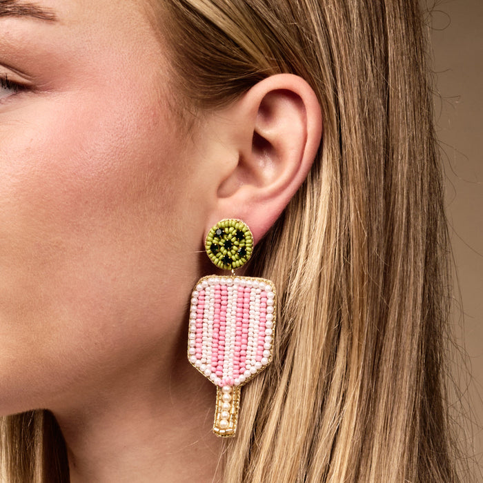 A close-up of a woman wearing a beaded earring shaped like a pink popsicle with a green circular stud. Only the left side of her face and ear are visible.