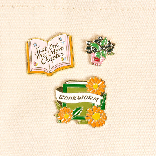 Three enamel pins: an open book with Just One More Chapter, a potted plant, and a green book with yellow flowers and a banner reading BOOKWORM, all on a cream fabric background.
