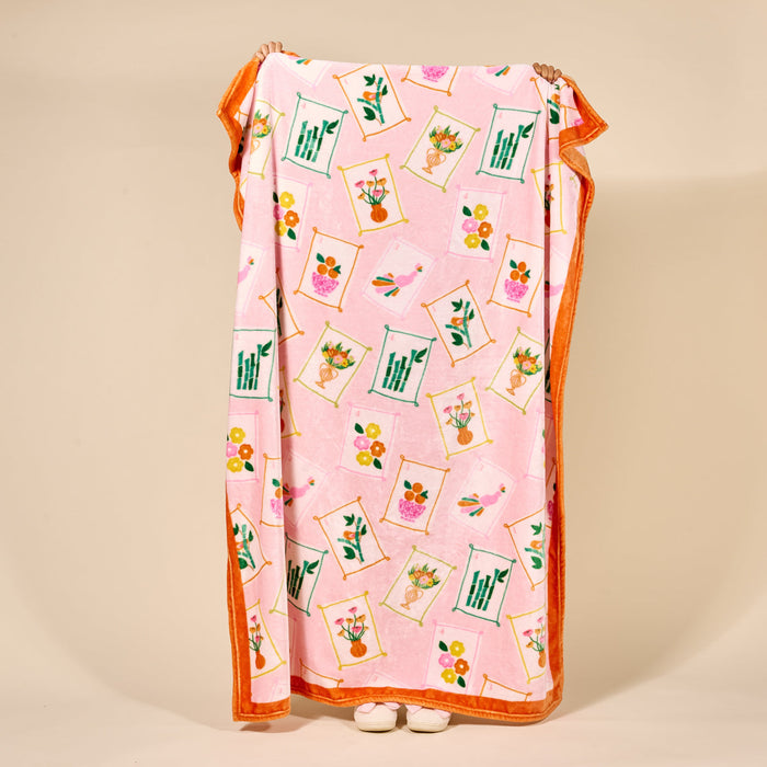 A person stands holding a pink blanket decorated with colorful drawings of flowers in vases and green plants. Only their hands and white shoes are visible behind the blanket.