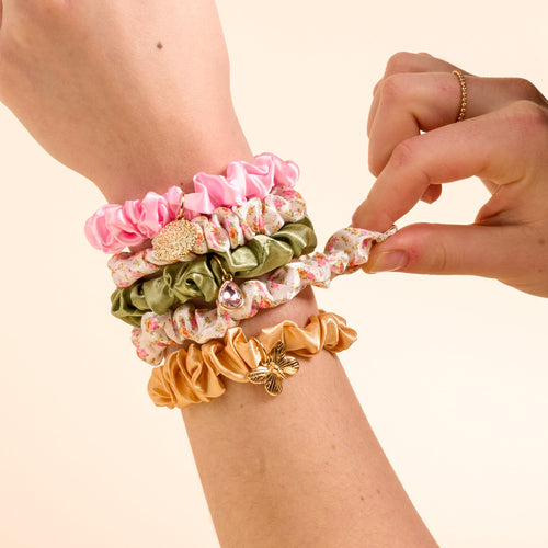 The Darling Effect's Endless Daydream Cream Scrunchie Charm Set features a satin scrunchie with hair charms, worn on the wrist and another being pulled gently by a finger.