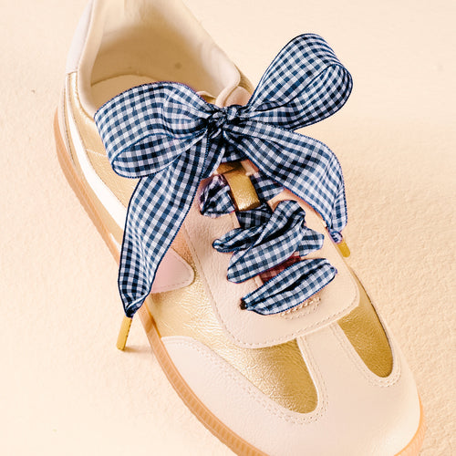 A gold and cream sneaker with The Darling Effect's Gingham Navy Step It Up Shoelaces, featuring a blue and white gingham pattern tied in a bow.