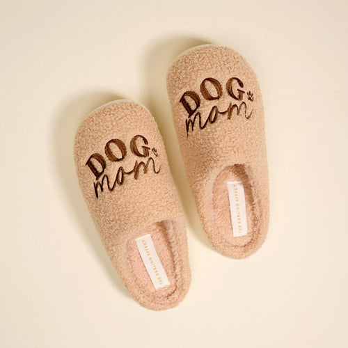A pair of fuzzy, light brown slippers with the words “DOG mom” embroidered in dark brown on the tops, shown on a light background.