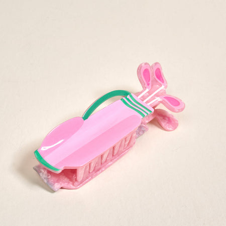 A pink hair clip shaped like a golf bag with golf clubs, featuring green and white details, is placed on a plain light surface.