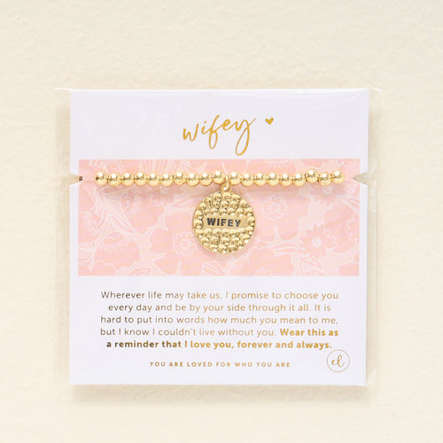 A gold beaded bracelet with a WIFEY charm is displayed on a card with a loving message in gold and gray text and a pink floral background.