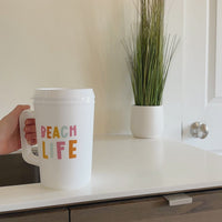 A hand holds the-darling-effect’s Beach Life 34 oz Mega Mug, featuring vibrant BEACH LIFE lettering, near a countertop with a white potted plant in the background—an ideal choice for staying hydrated thanks to its spacious 34 oz capacity.