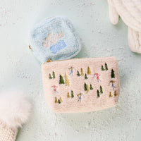 Two fuzzy Square Aprés Ski Teddy Pouches by The Darling Effect rest on a light surface. The blue sherpa pouch features “Après Ski” embroidery, while the beige pouch is adorned with small embroidered skiers and trees. Snow-like glitter and white knit accessories surround them, enhancing the cozy après-ski vibe.