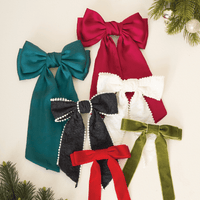 The Darling Effect’s Black Pearl Hair Bow, an elegant fabric accessory, is featured among red, teal, white, and green velvet bows on a light background with Christmas greenery accents.