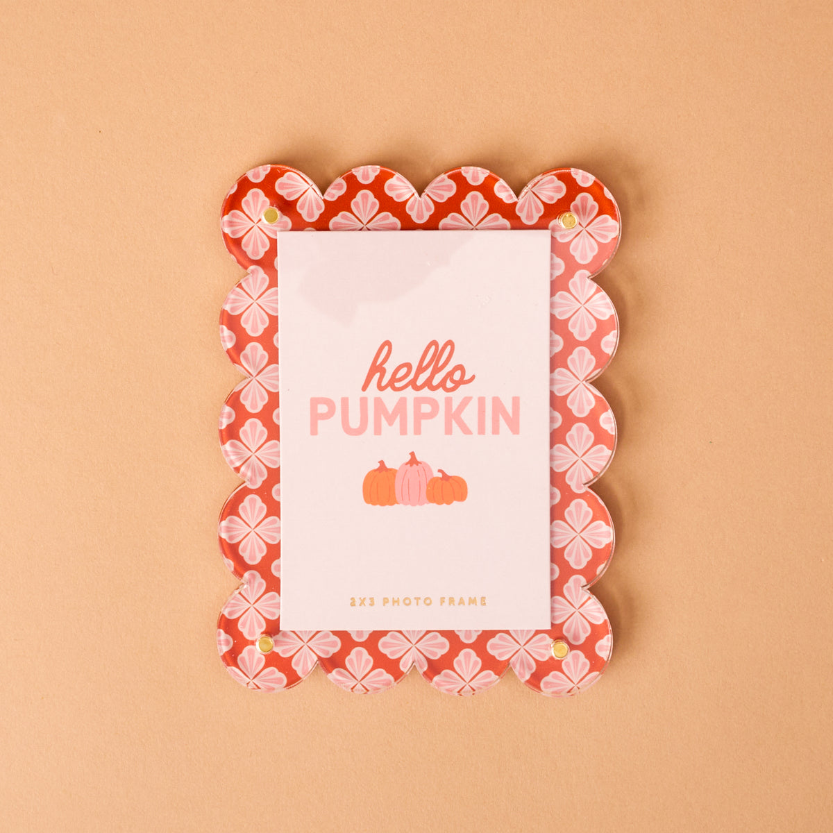 A scalloped, pink and red Sienna Spice Mini Acrylic Picture Frame Magnet by The Darling Effect features floral patterns and holds a card that says “hello PUMPKIN” with three pumpkins, set against a solid peach background.