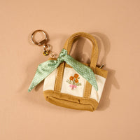 The Honey Blossom Tiny Tote Bag Charm by The Darling Effect is a small beige and white mini tote bag keychain, adorned with an embroidered flower design and a green polka-dot scarf tied to one handle. It features a convenient zipper compartment and is displayed on a tan background.