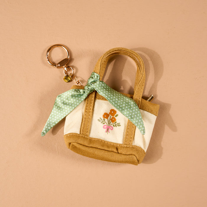 The Honey Blossom Tiny Tote Bag Charm by The Darling Effect is a small beige and white mini tote bag keychain, adorned with an embroidered flower design and a green polka-dot scarf tied to one handle. It features a convenient zipper compartment and is displayed on a tan background.