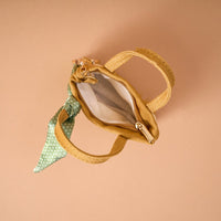 The Honey Blossom Tiny Tote Bag Charm by The Darling Effect—a small tan handbag featuring a zipper compartment, white interior, and a green polka dot scarf tied to one handle—is displayed on a beige background. This charming mini tote bag charm brings playful elegance to any accessory collection.