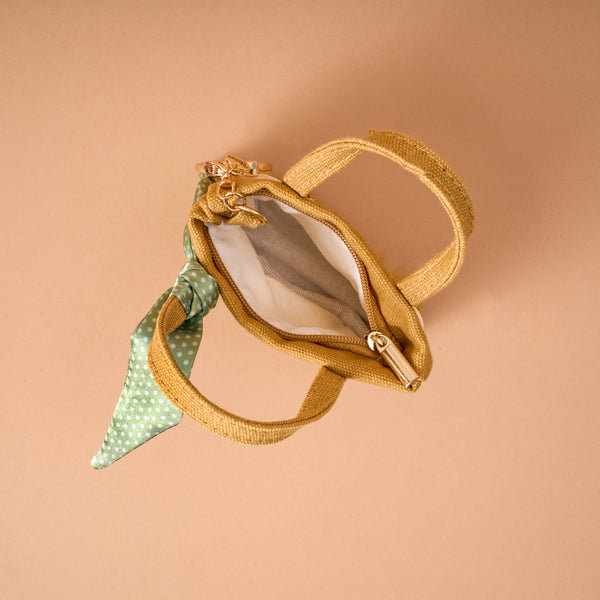 The Honey Blossom Tiny Tote Bag Charm by The Darling Effect—a small tan handbag featuring a zipper compartment, white interior, and a green polka dot scarf tied to one handle—is displayed on a beige background. This charming mini tote bag charm brings playful elegance to any accessory collection.