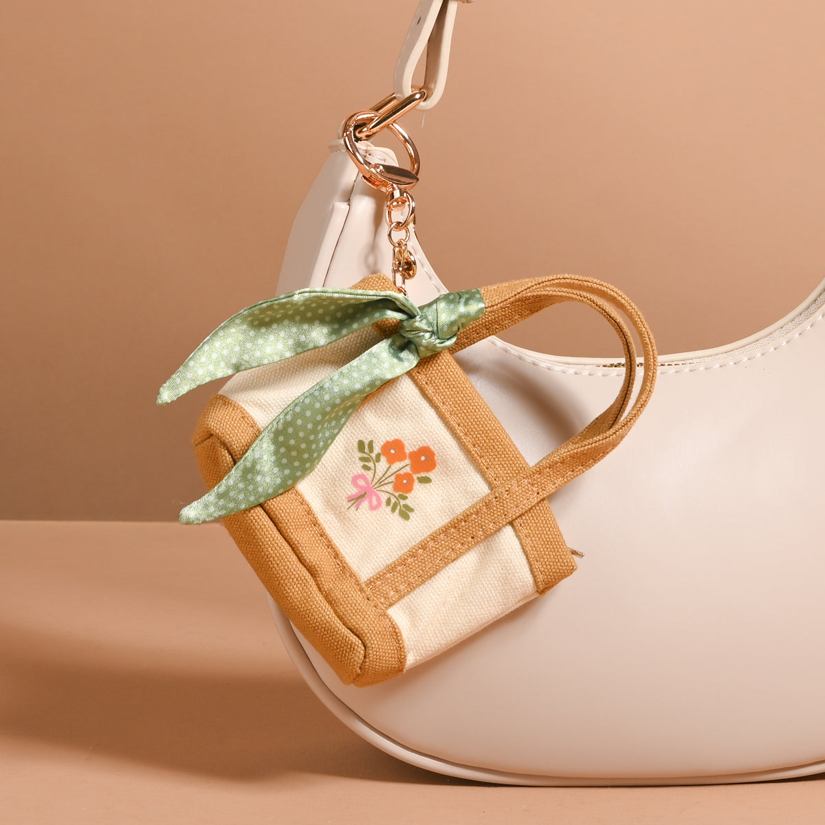 The Honey Blossom Tiny Tote Bag Charm by The Darling Effect, featuring beige and tan tones with embroidered flowers and a green polka dot bow, is styled as a mini tote bag charm attached to a light cream-colored handbag on a neutral background.